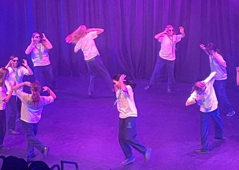 Pupils dancing in a circle wearing white shirts with green ties and sunglasses.